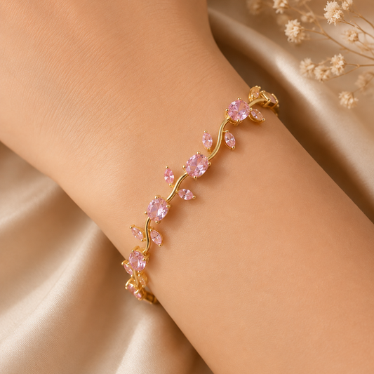 A gold vine bracelet with pink faceted crystal leaves, worn on a model's wrist against a beige satin background with dried flowers.