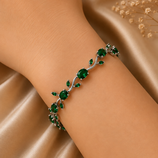 A silver-toned vine bracelet adorned with round and teardrop-cut emerald green gemstones, shown worn on a person's wrist against a draped champagne satin background with dried flowers.