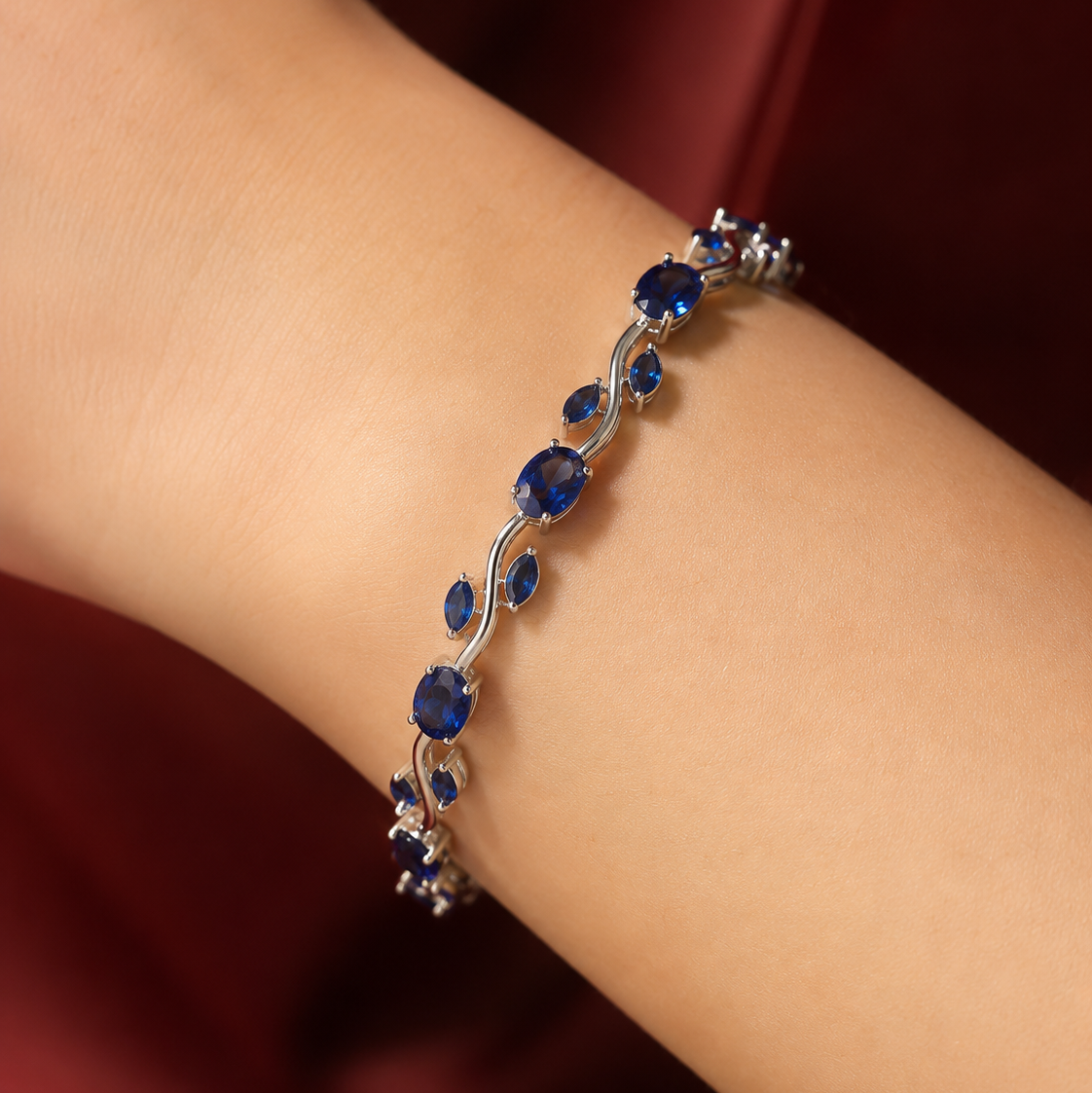 A close-up of a silver-toned bracelet with deep blue oval and teardrop gemstones, worn on a person's wrist against a dark red satin background.