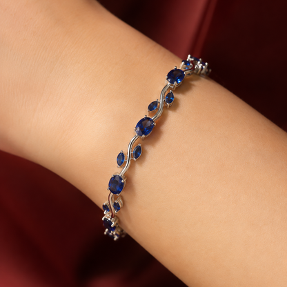 A close-up of a silver-toned bracelet with deep blue oval and teardrop gemstones, worn on a person's wrist against a dark red satin background.