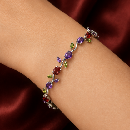 A silver-toned bracelet with colorful gemstones arranged in a vine pattern, worn on a person's wrist against a dark red satin background.