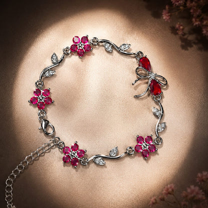 A silver-toned bracelet decorated with pink gemstone flowers, leaf motifs, and a butterfly charm, displayed on a warm brown background with soft floral accents.