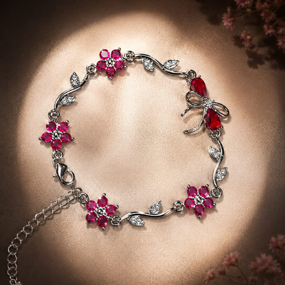 A silver-toned bracelet decorated with pink gemstone flowers, leaf motifs, and a butterfly charm, displayed on a warm brown background with soft floral accents.