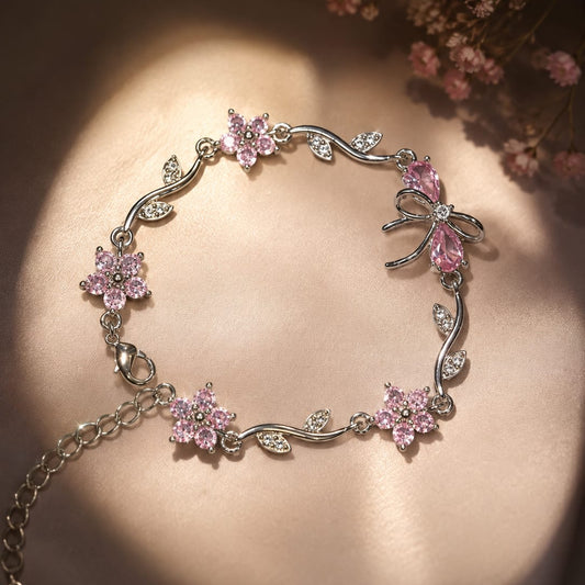 A silver-toned floral bracelet with pink gemstone flowers and bow details, displayed on a warm beige satin surface with dried pink flowers in the corner.