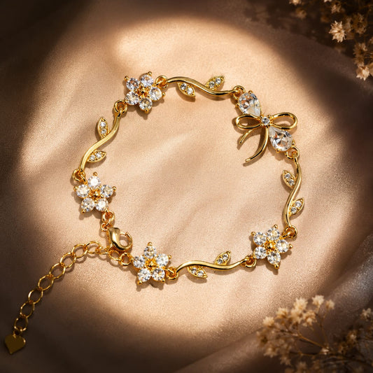 A gold bracelet with floral and vine motifs set with clear crystals, displayed on a draped beige satin fabric with warm sunlight and dried flowers in the corners.