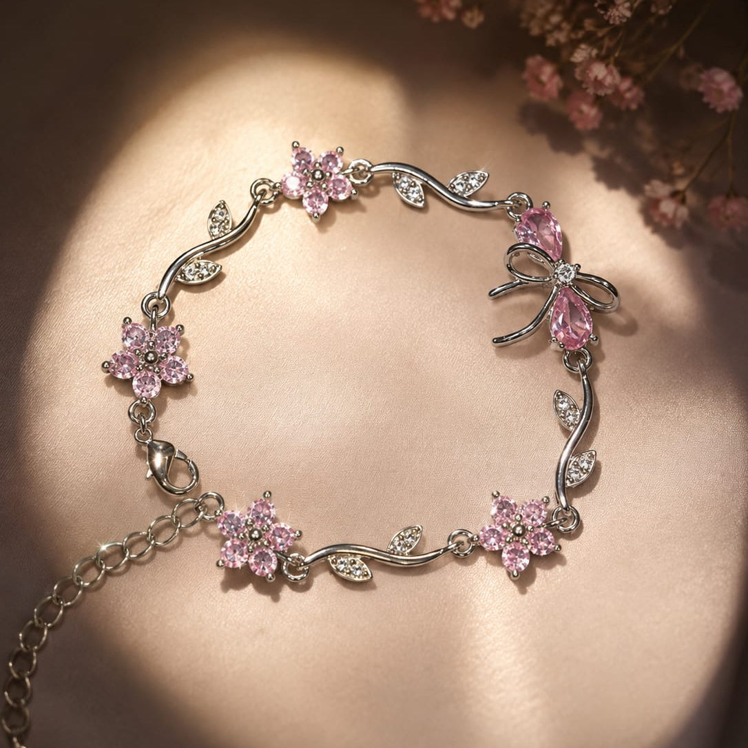 A silver-toned floral bracelet with pink gemstone flowers and bow details, displayed on a warm beige satin surface with dried pink flowers in the corner.