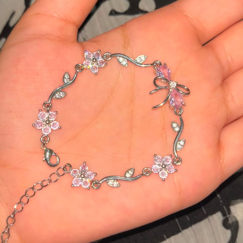 A silver-toned bracelet with pink crystal flowers and bow details, displayed on an open hand.